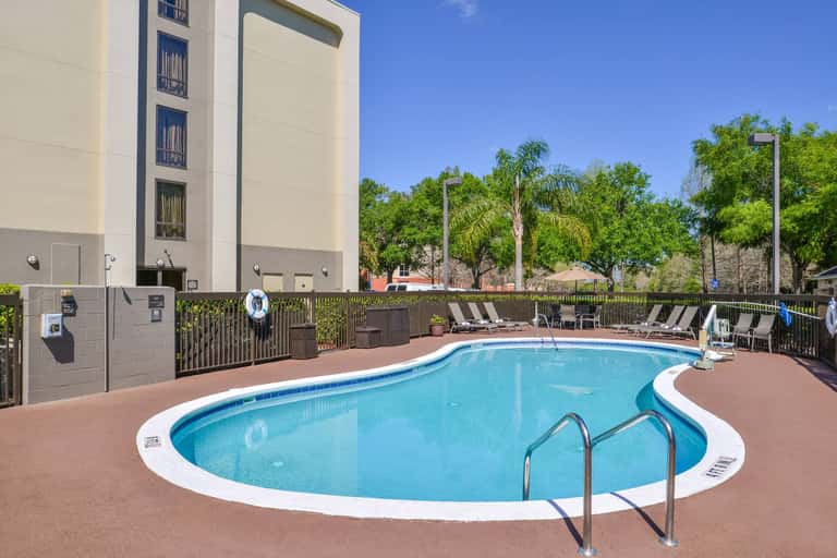 Hotel pool with blue water, white trim, ladder, lounge chairs, and modern building adjacent