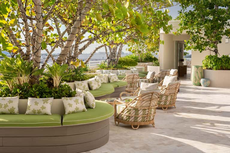 Shaded rooftop terrace with curved green seating, rattan chairs, tropical plants, and ocean views