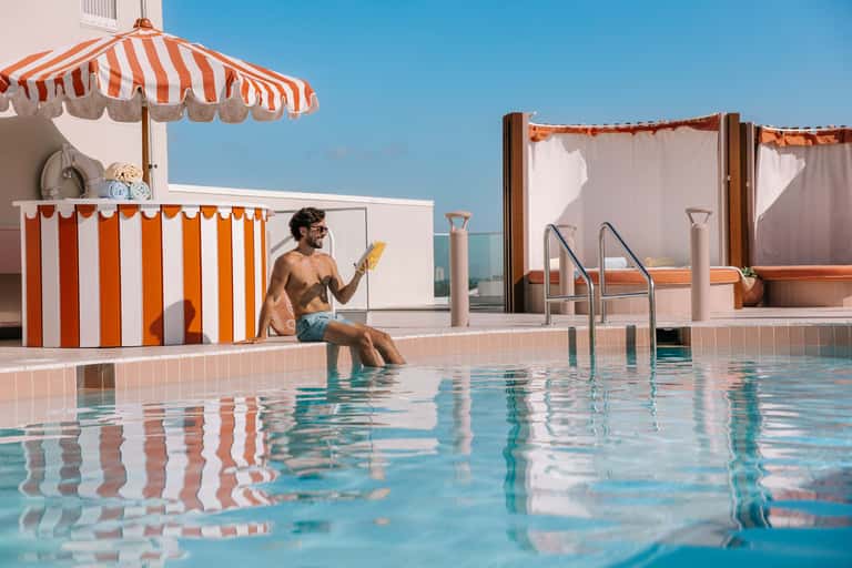 Man in pool at retro-style resort with red and white striped umbrellas and beach cabana