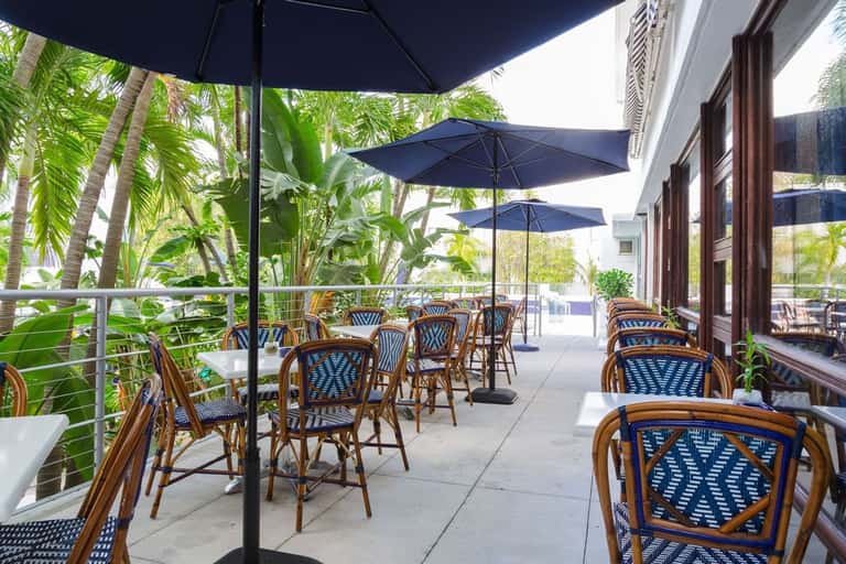 Outdoor dining patio with rattan chairs, blue patterned seat cushions, black umbrellas, tropical plants, and glass building facade