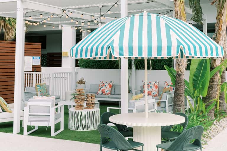 Tropical resort patio with turquoise striped umbrella, white furniture, and palm trees under string lights