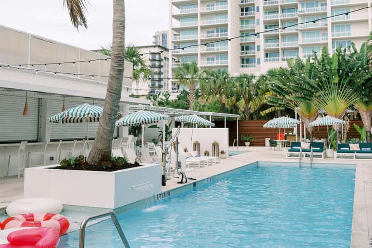 Resort pool surrounded by green-striped umbrellas, loungers, and multi-story hotel building