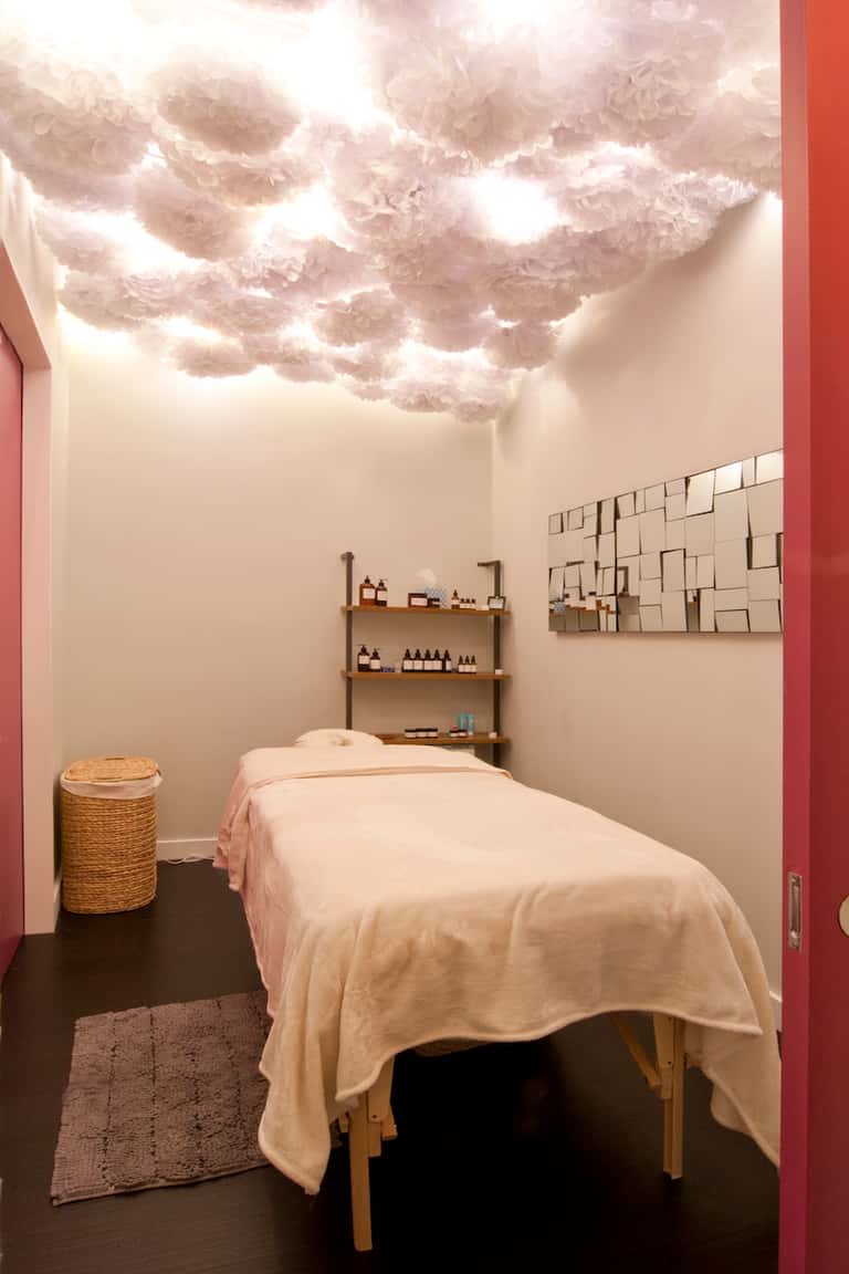 Spa treatment room with massage bed, fluffy cloud ceiling light installation, wooden shelving with products, and neutral décor