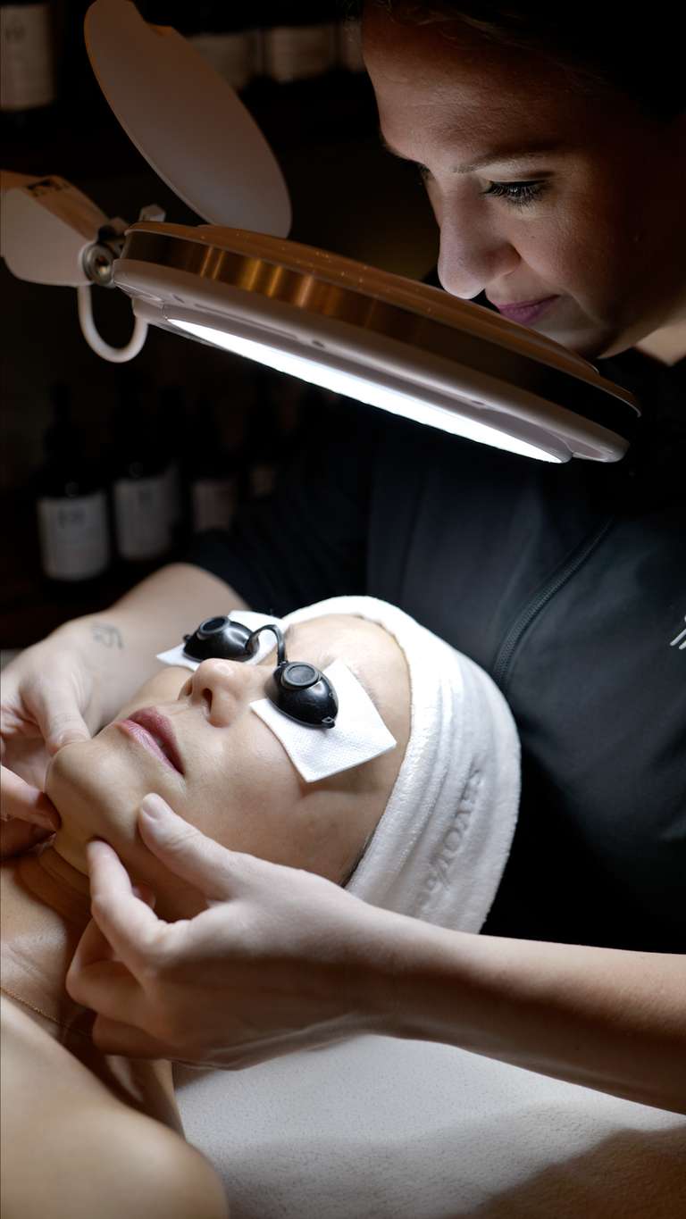 Esthetician applying spa facial treatment with handheld device while client relaxes wearing headband and eye patches