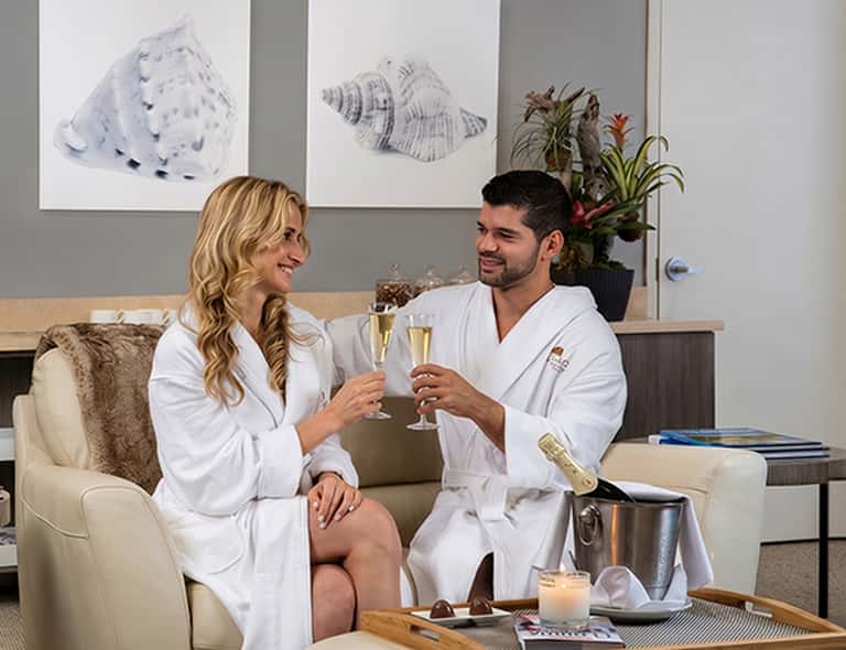 Couple in white robes clinking champagne flutes in a spa suite with shell artwork and plants
