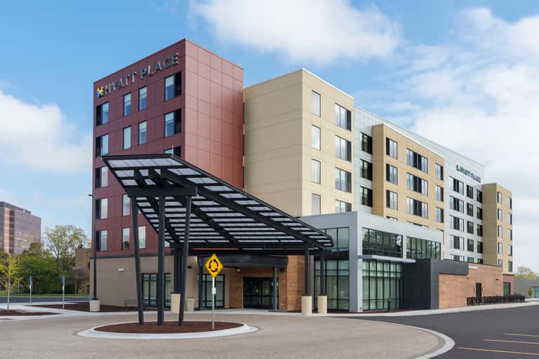 Modern Hyatt Place hotel exterior with burgundy tower, beige wings, black pergola entrance canopy, and circular courtyard