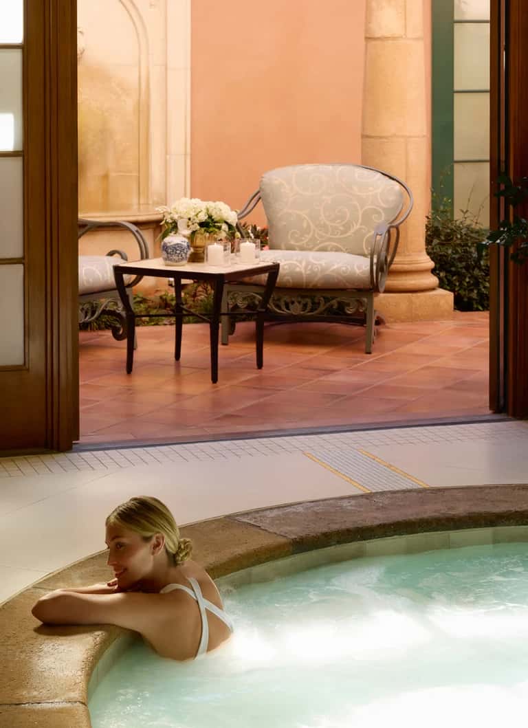 Woman relaxing in hot spring spa pool with courtyard seating area and decorative flowers in background