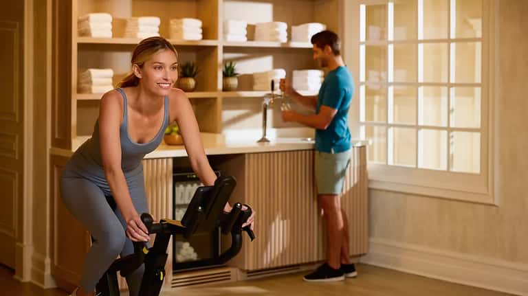 Woman on stationary bike in home gym with man getting water, wooden shelving with towels, modern aesthetic