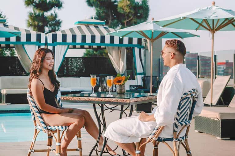 Couple enjoying drinks at a poolside table under turquoise umbrellas and striped cabanas.
