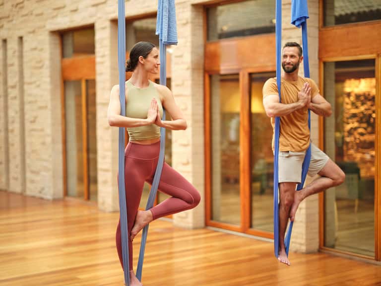 Man and woman performing aerial yoga poses using blue silk hammocks suspended in a modern courtyard with stone columns