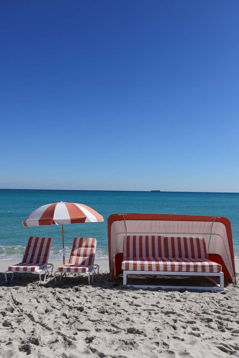 Red and white striped beach loungers and tent with umbrella on sandy beach facing turquoise ocean