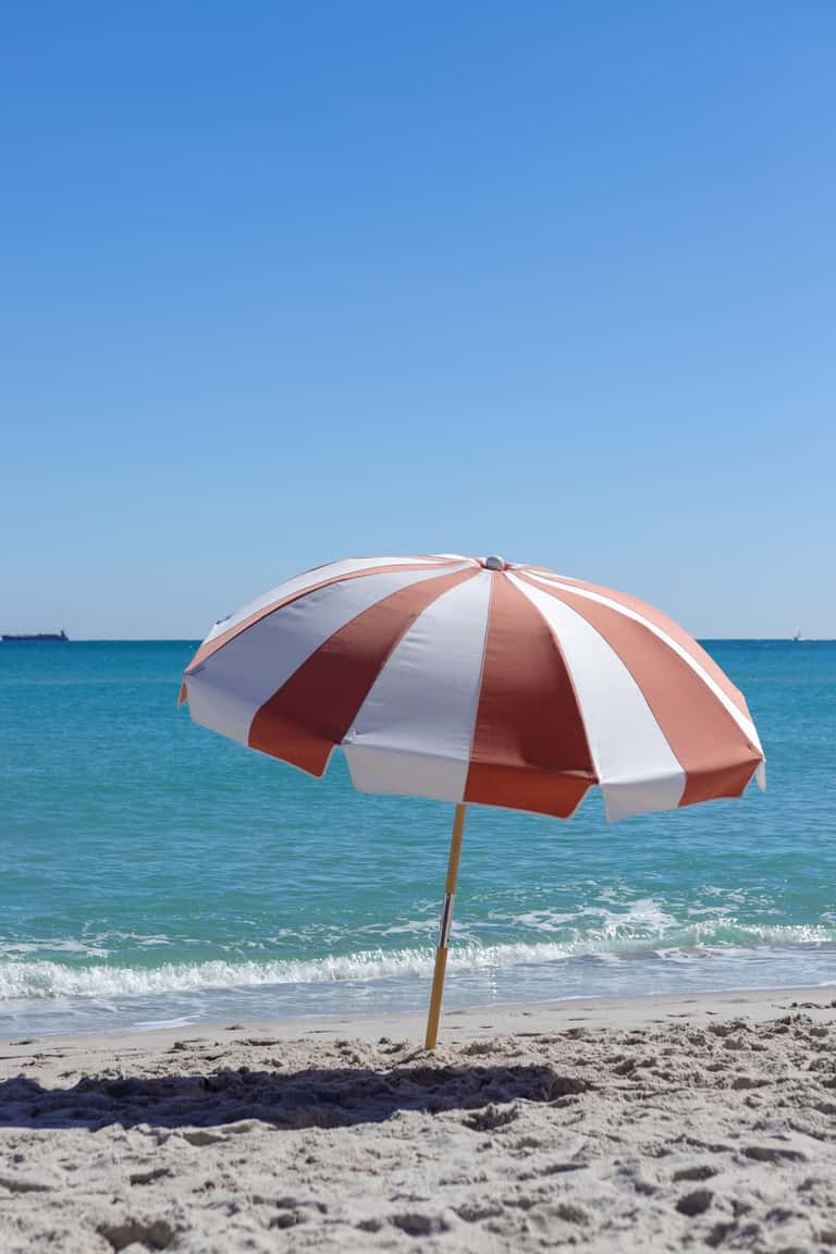 Red and white striped beach umbrella planted in sand overlooking ocean waves