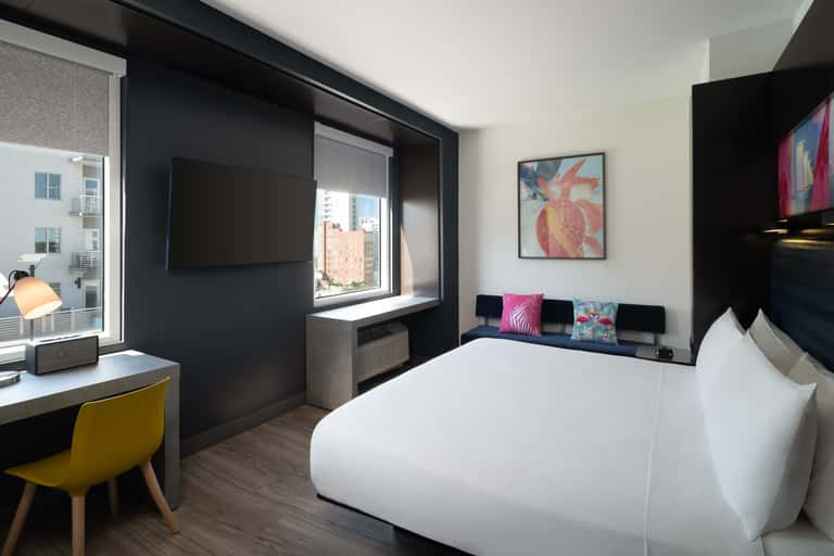 Hotel bedroom with queen bed, black accent wall, yellow desk chair, and city skyline views