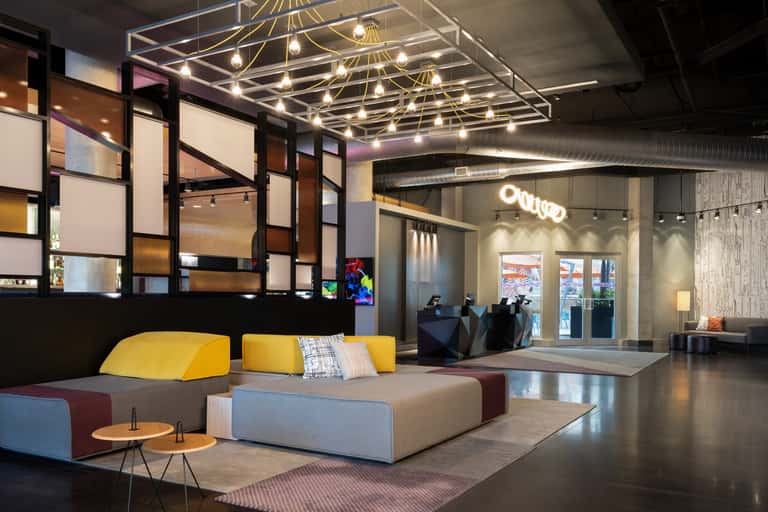 Contemporary hotel lobby with modular seating, yellow and blue upholstery, brass ceiling fixture, and geometric wall partitions