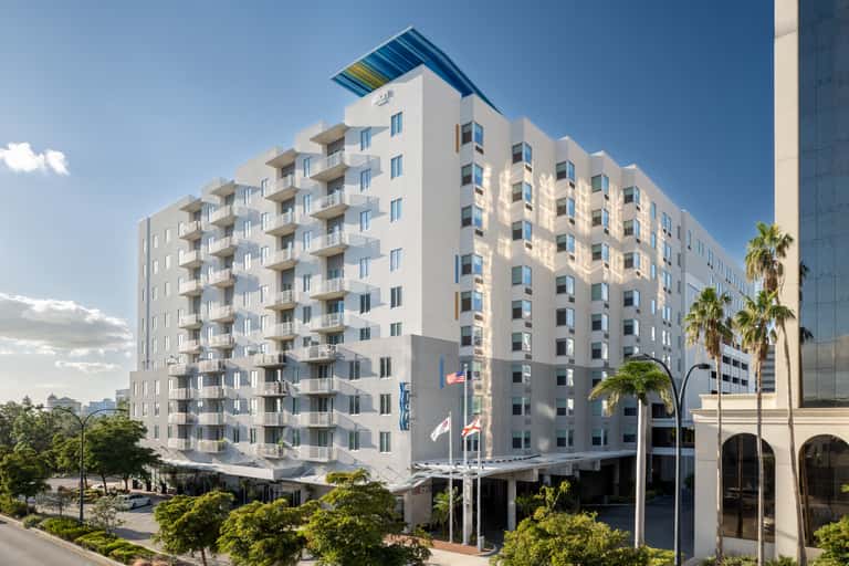 Eight-story white residential building with balconies, colorful roof accent, palm trees, and ground-level retail spaces