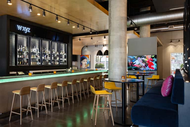 Hotel bar and lounge with illuminated bar counter, yellow chairs, TV screens, and contemporary design elements