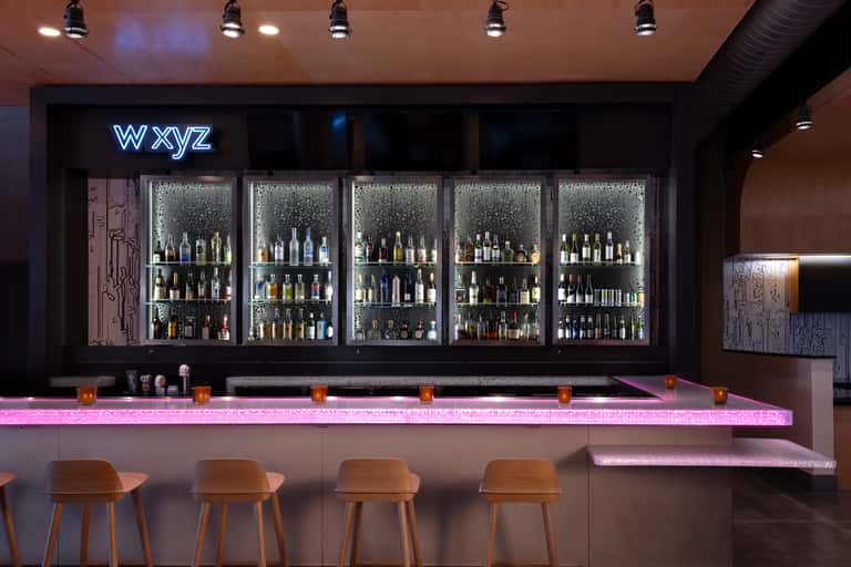 Modern bar interior with neon 'wxyz' sign, illuminated pink counter, backlit bottle display, and wooden bar stools