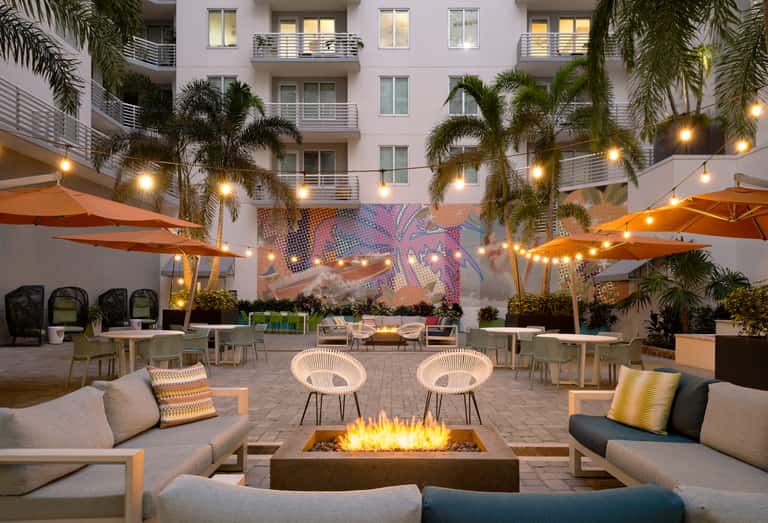 Courtyard gathering space with fire pit, lounge seating, palm trees, orange umbrellas, and colorful wall mural