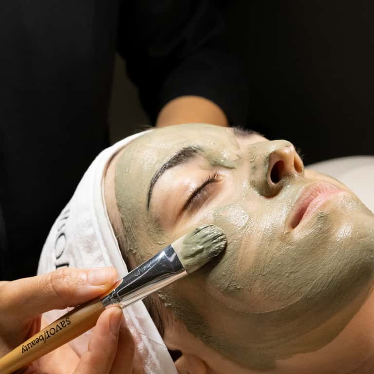 Esthetician applying green clay mask to client's face with professional brush