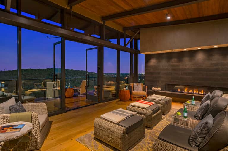 Modern lounge with woven furniture, fireplace, and expansive windows overlooking hillside