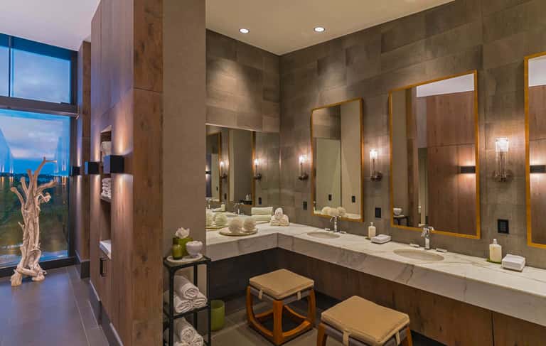 Spa bathroom with dual vanities, illuminated mirrors, and marble countertops overlooking sunset