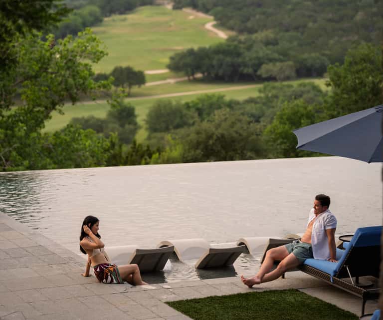 Couple relaxing by modern infinity pool overlooking rolling green landscape and hills