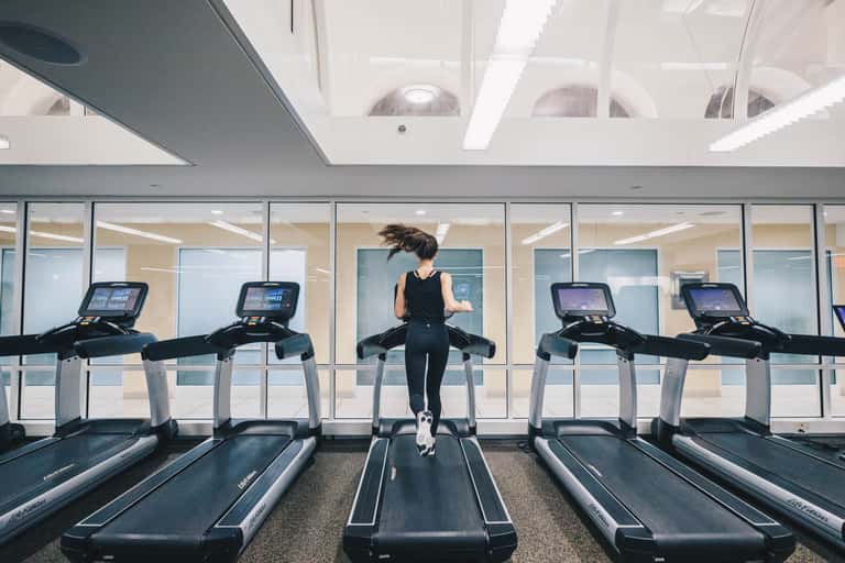 Woman jogging on treadmill in modern, bright gym with floor-to-ceiling windows and multiple cardio machines