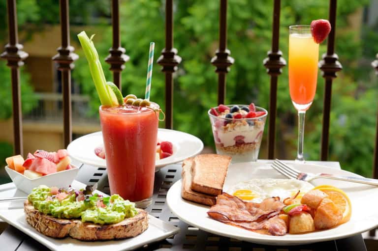 Outdoor brunch spread with Bloody Mary cocktail, avocado toast, eggs, bacon, pastries, and fresh fruit on white plates