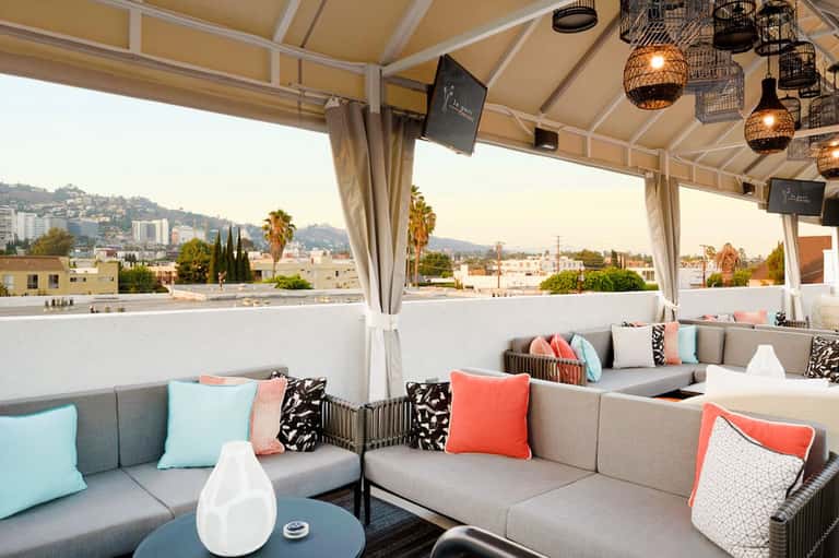 Covered rooftop lounge with gray seating, colorful pillows, and hillside city views