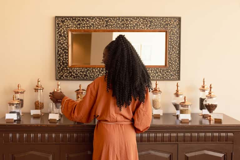 Woman in orange robe viewing mirror above vanity with decorative containers and toiletries