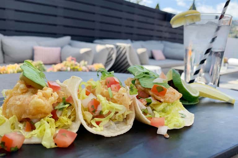 Three fish tacos with fresh garnish on dark plate overlooking resort lounge area