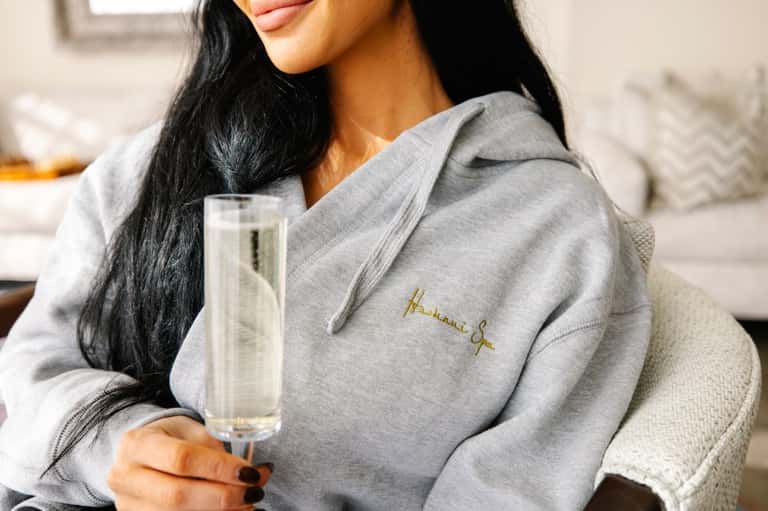 Woman in gray hoodie holding champagne flute, relaxing on neutral-toned seating