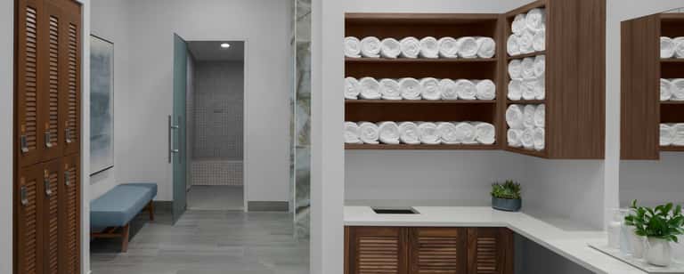 Modern spa bathroom with wood cabinetry, white rolled towels on shelves, shower enclosure, and white countertop with plants