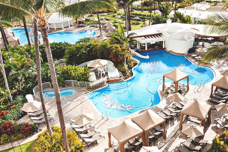 Aerial view of resort with curved blue pools, palm trees, lounge chairs, and umbrellas surrounded by manicured grounds