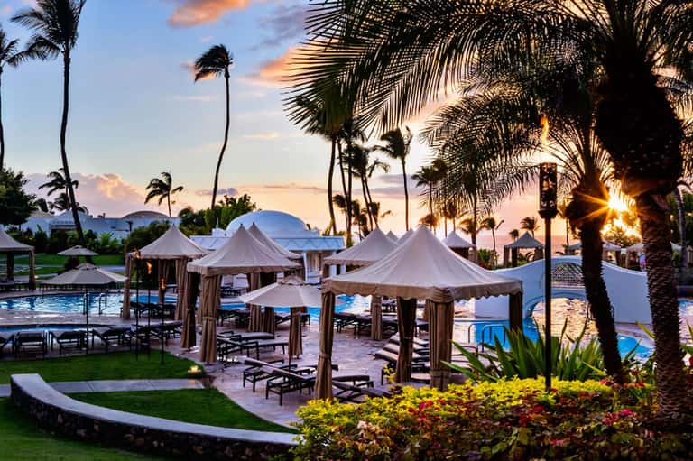Beachfront resort at dusk with white cabanas, palm trees, and illuminated pool area