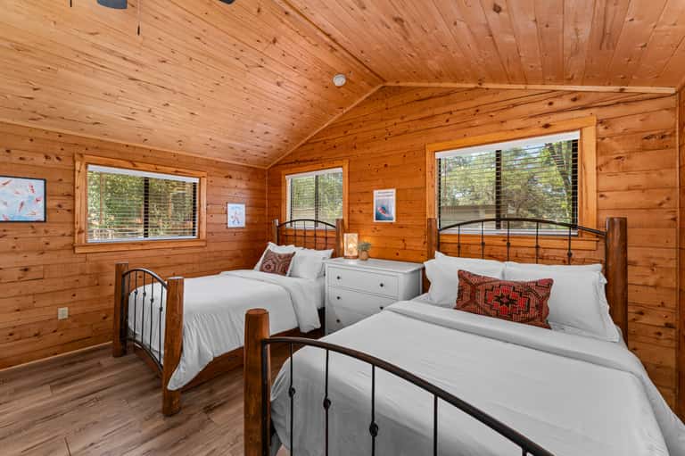 Spacious wooden cabin bedroom with two metal-frame beds, white linens, red pillows, and large windows overlooking trees