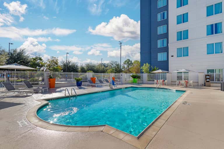 Outdoor pool area with turquoise water, lounge chairs, white railings, and modern hotel building backdrop