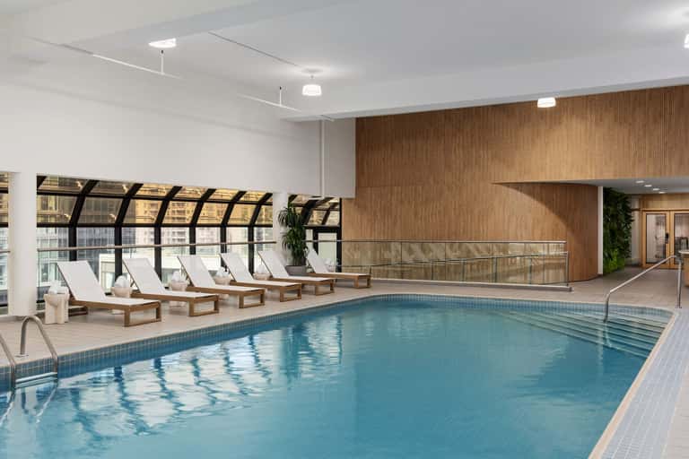 Indoor pool with wooden loungers, white cushions, and floor-to-ceiling windows in luxury facility