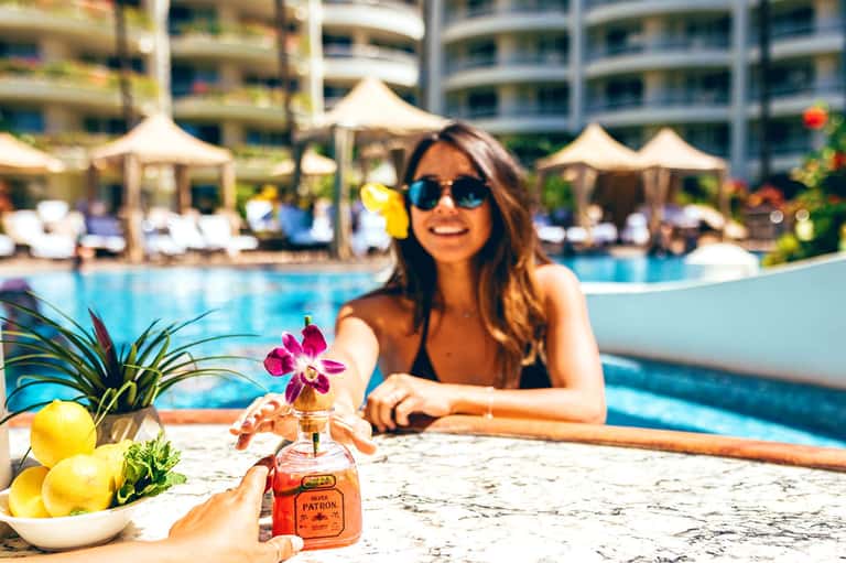 Woman in sunglasses at resort pool holding Patrón tequila bottle with lemons and flowers