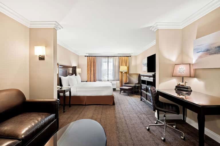 Spacious hotel suite with king bed, seating area, fireplace, dark wood furniture, and work desk