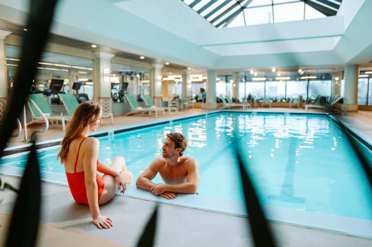 Man and woman by indoor lap pool with lounge chairs in bright, modern facility