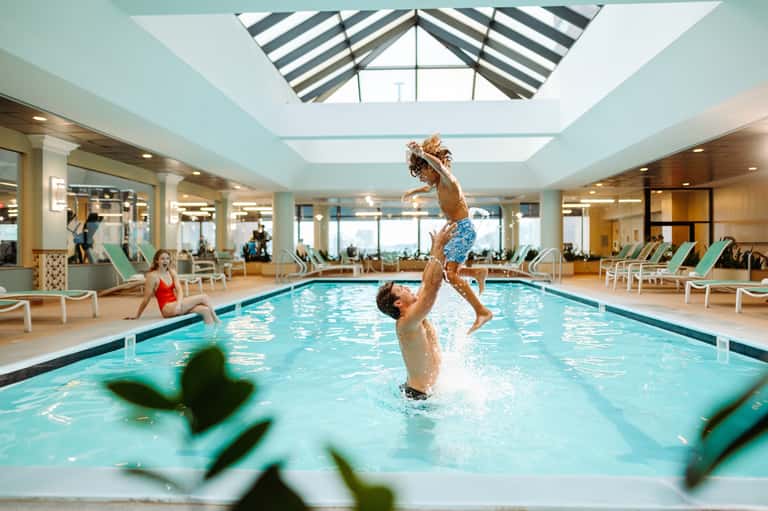 Father tossing child in air above indoor pool with skylights and lounge seating