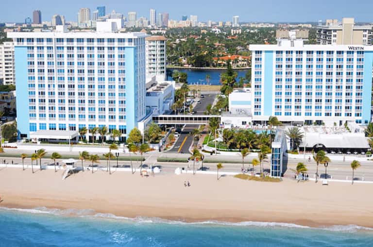 Beachfront resort with bright blue twin towers, sandy beach, ocean waves, and downtown skyline in background
