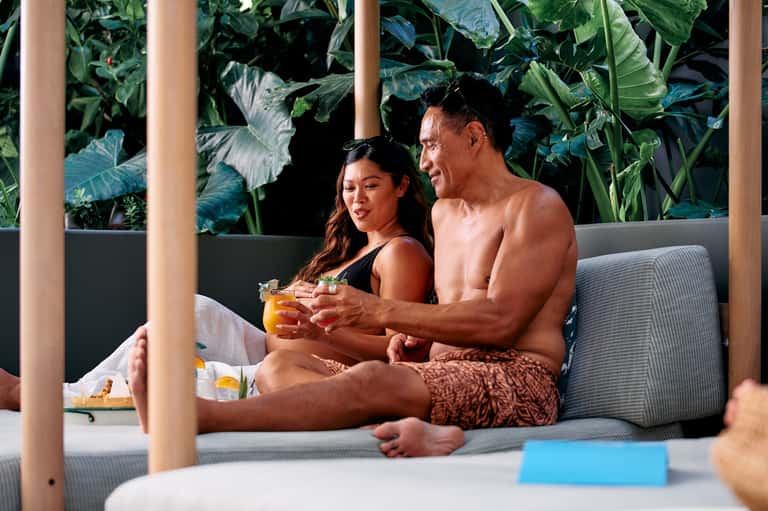 Couple relaxing on a lounge chair with tropical foliage backdrop, holding cocktails at resort