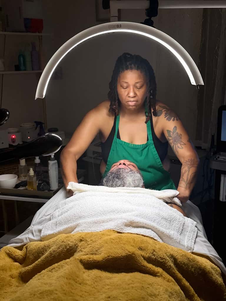Esthetician in green apron performing facial treatment under ring light on client with face mask