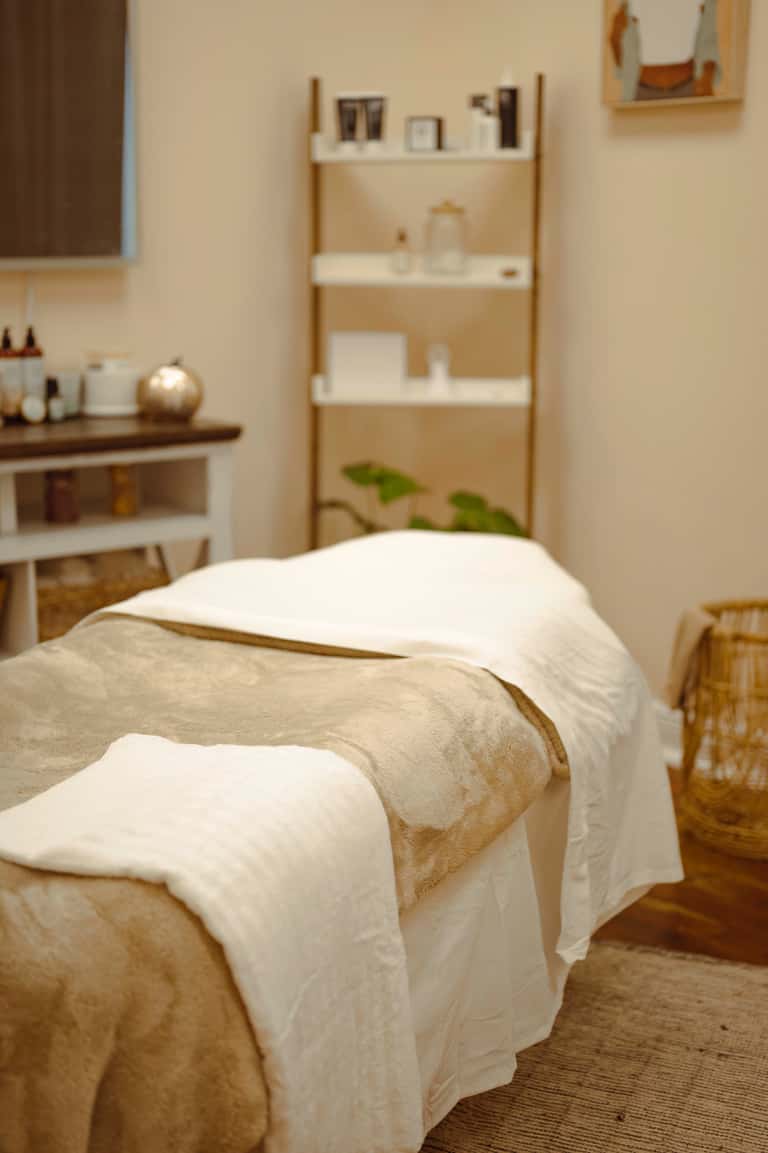 Minimalist spa treatment room with beige walls, wooden shelving, vanity, and massage bed