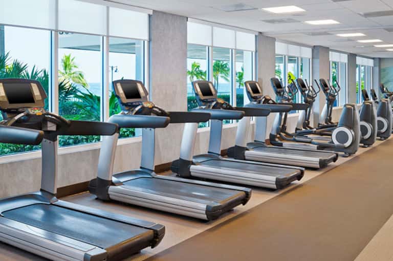 Modern fitness center with treadmills and elliptical machines along floor-to-ceiling windows overlooking tropical gardens