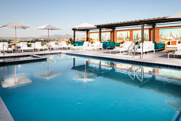 Modern rooftop pool with turquoise water, white loungers, orange building, and desert mountain views