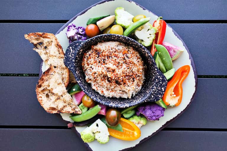 Hummus dip in black bowl surrounded by fresh vegetables and toasted bread on white plate