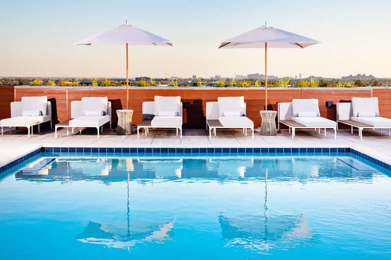 Resort rooftop pool with blue water, white loungers, umbrellas, and wooden privacy wall overlooking landscape